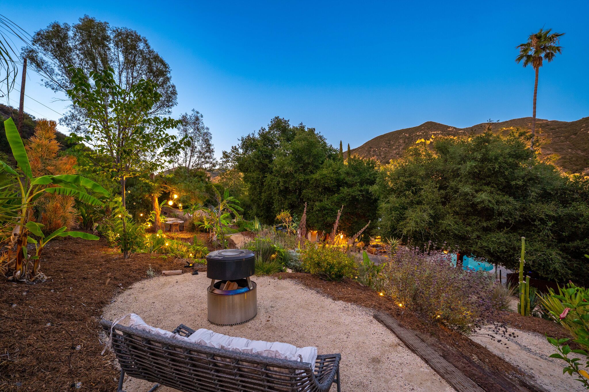 Fire table burning at dusk on a gravel terrace with lit gardens and canyon ridgelines beyond