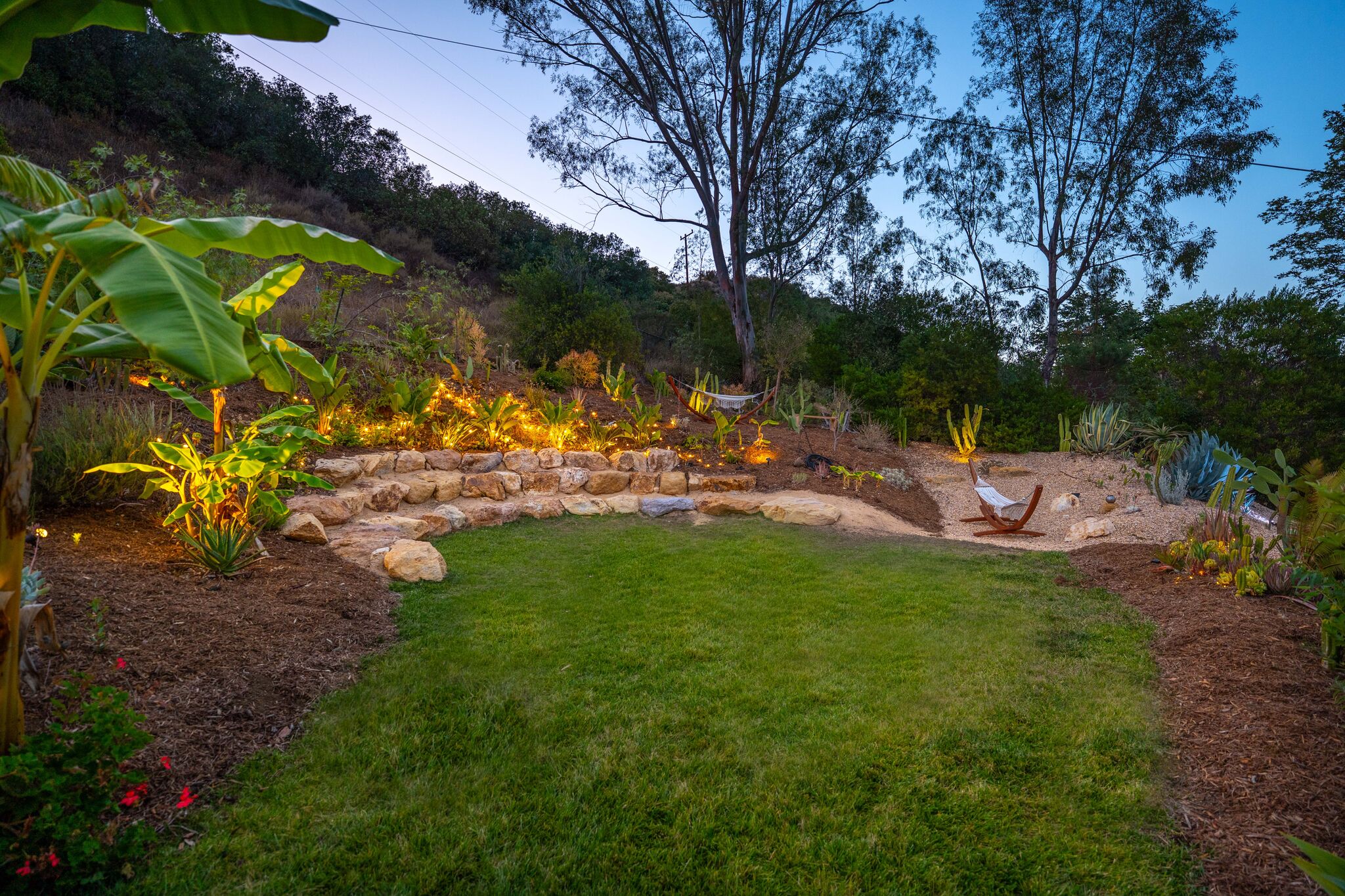 Lawn at dusk edged by a boulder terrace with glowing garden lights and a hanging chair