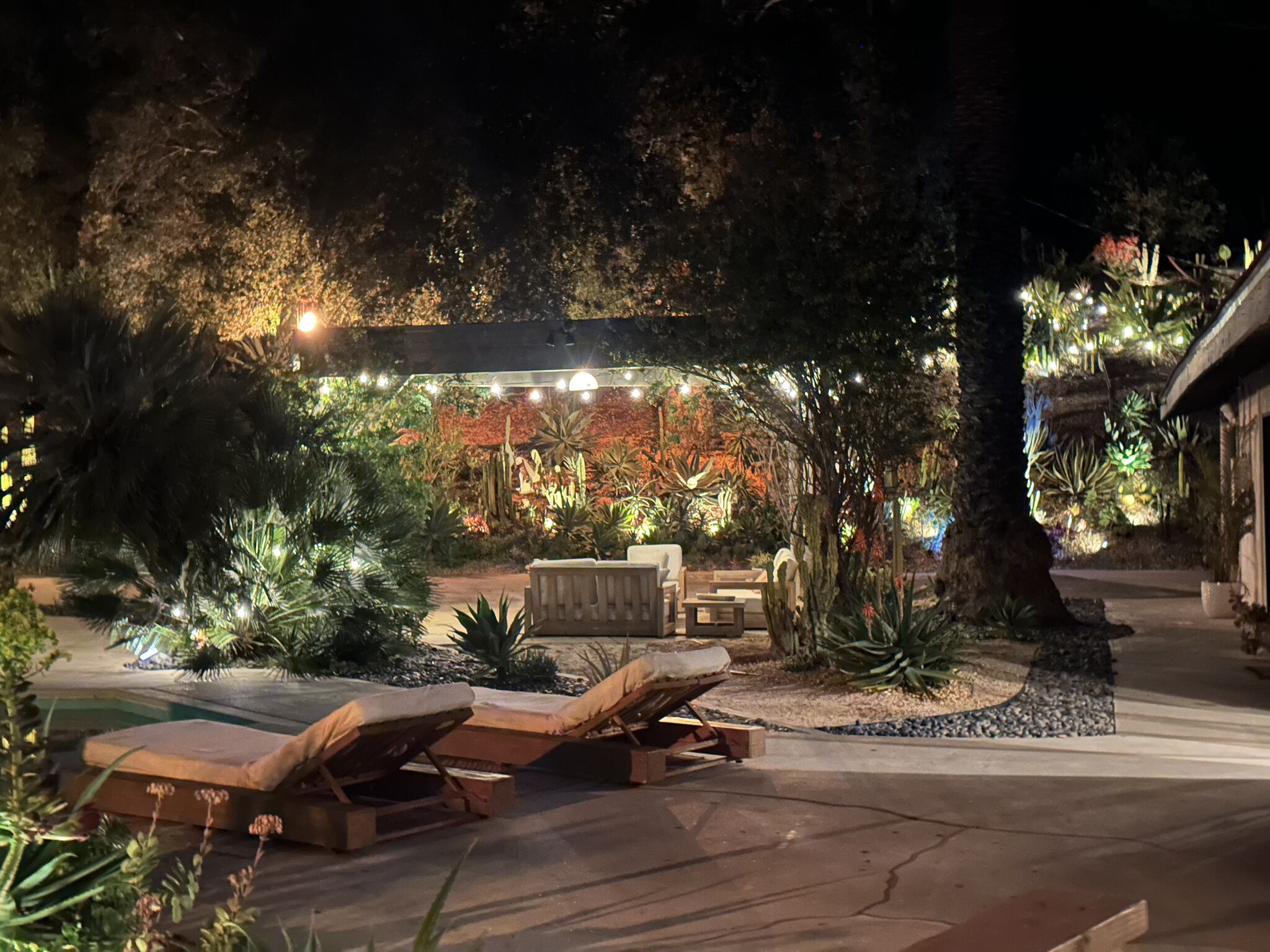 Pool deck loungers at night beneath string lights with the lit gardens glowing behind